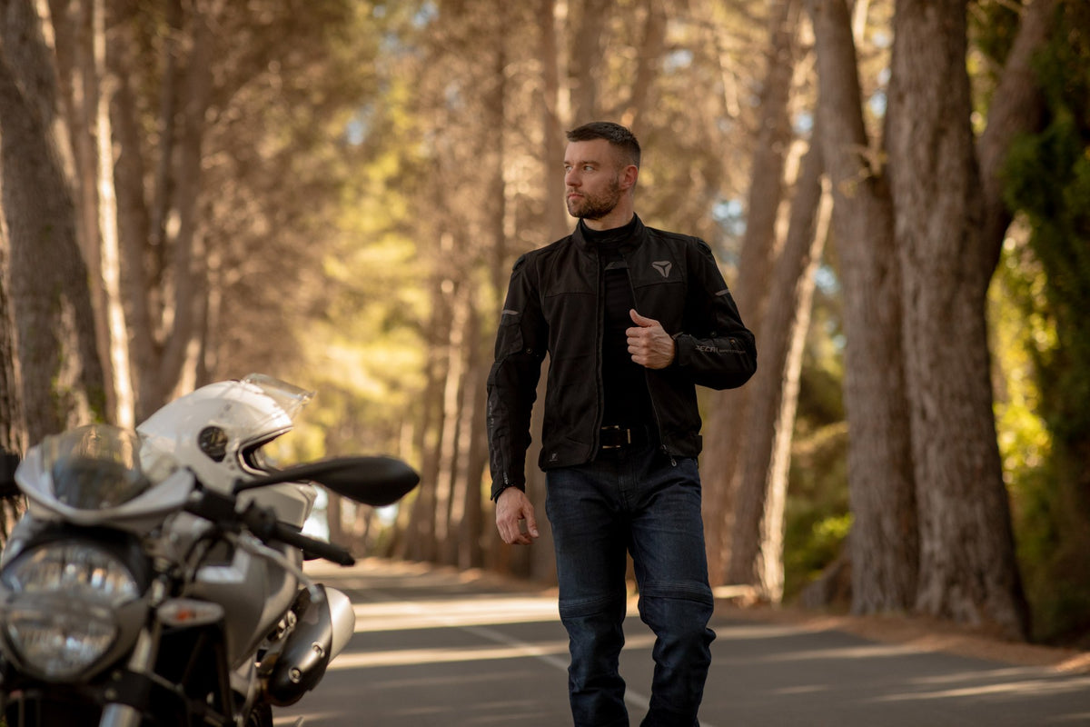 Man bär Seca Textil Mc-Jacka Venti Pro Svart vid en landsväg med motorcykel i bakgrunden. Perfekt för stil och skydd vid motorcykelkörning.